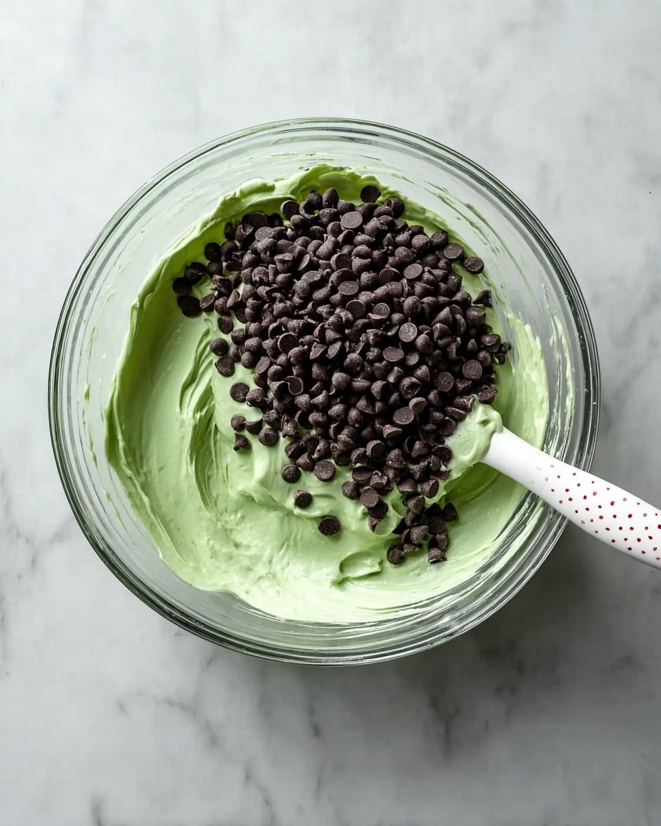 A clear glass bowl sits on a white marbled surface filled with light green creamy mixture that looks smooth and fluffy. On top of the green cream, there is a thick layer of small dark chocolate chips scattered mostly on the right side, sitting like a mound. Inside the bowl is a white spatula with a white handle dotted with tiny red spots, partially submerged in the green cream near the chocolate chips. The scene is bright and clear, showing all the textures and colors clearly photo taken with an iphone --ar 4:5 --v 7