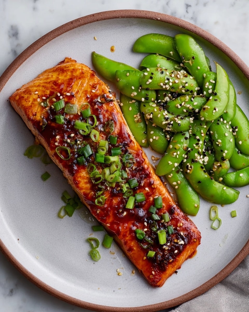 A piece of cooked salmon with a shiny dark orange glaze sits diagonally on a white plate. It is topped with small chopped green onions and sprinkled with white and black sesame seeds. On the right side of the plate, there is a pile of bright green edamame pods with some sesame seeds on top. The plate rests on a white marbled surface. Photo taken with an iphone --ar 4:5 --v 7
