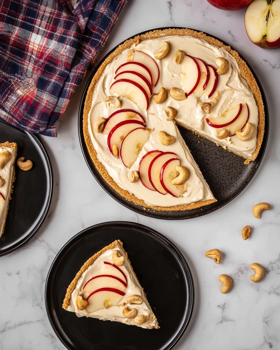 A large round tart with a golden crust forms the base layer, topped evenly with a thick layer of creamy, light beige frosting. On top of the frosting, there are thin slices of red apple arranged in pairs, slightly overlapping, each with a fresh and shiny appearance. Small clusters of light brown cashew nuts are scattered across the tart surface, adding a textured, crunchy look. Two triangular slices of the tart are placed on separate glossy black plates below and to the side of the main tart, showing the same layering of crust, frosting, apple slices, and nuts. The setup is on a white marbled surface with a few loose cashews and apple slices spread around, along with a red and blue checked cloth near the edge. Photo taken with an iphone --ar 4:5 --v 7