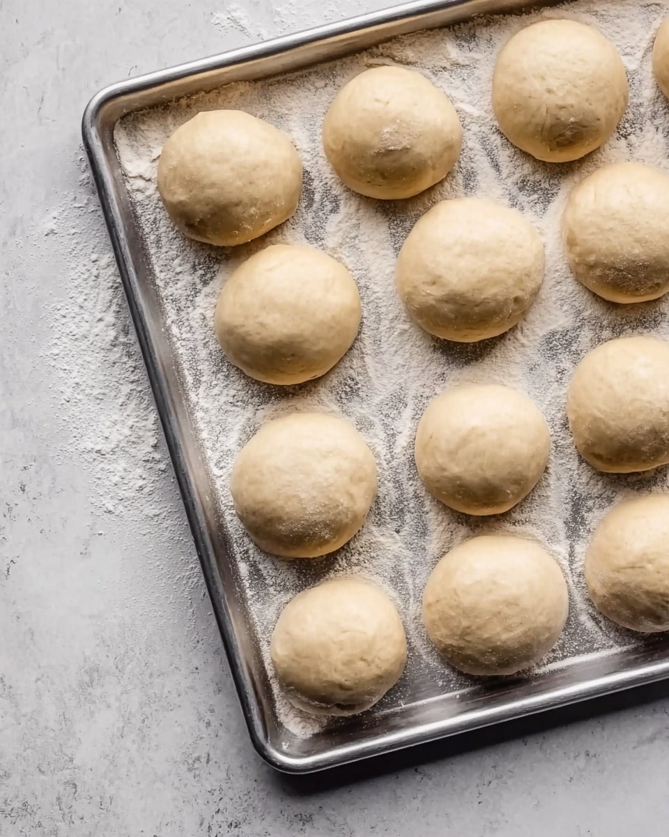 The image shows a metal baking tray filled with thirteen round dough balls arranged neatly in three rows on a surface lightly dusted with flour. Each dough ball is smooth and pale beige in color, with a slightly shiny texture, and they are evenly spaced on the tray. The tray sits on a white marbled textured surface, adding a clean and simple background to the scene. The dough balls appear soft and ready to be baked. photo taken with an iphone --ar 4:5 --v 7