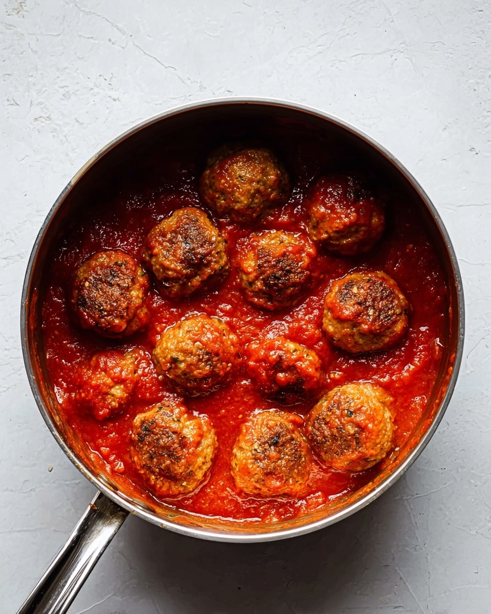 A stainless steel pot filled with twelve round, browned meatballs sitting in a thick, bright red tomato sauce. The meatballs have a slightly rough texture with some darker spots, and the sauce is smooth with visible small chunks of tomato. The pot handle extends to the left and the pot is placed on a white marbled textured surface. photo taken with an iphone --ar 4:5 --v 7