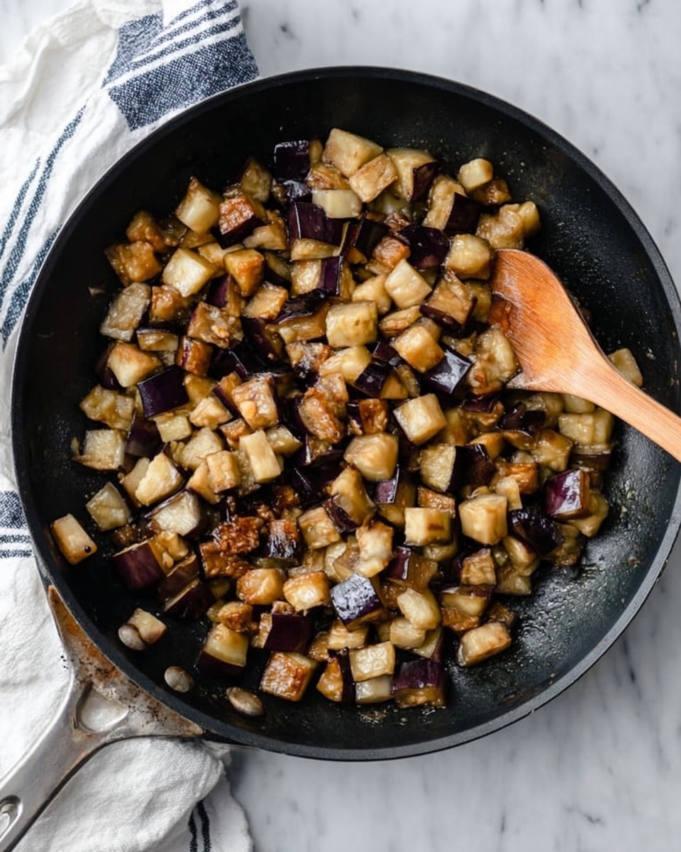 A black pan filled with small cubes of cooked eggplant in different shades of light brown, cream, and deep purple, showing a slightly soft and shiny texture from cooking. A wooden spoon is resting on the right side of the pan, partially covered by the eggplant cubes. The pan is sitting on a white marbled surface with a white and blue striped cloth partially under the left side. photo taken with an iphone --ar 4:5 --v 7