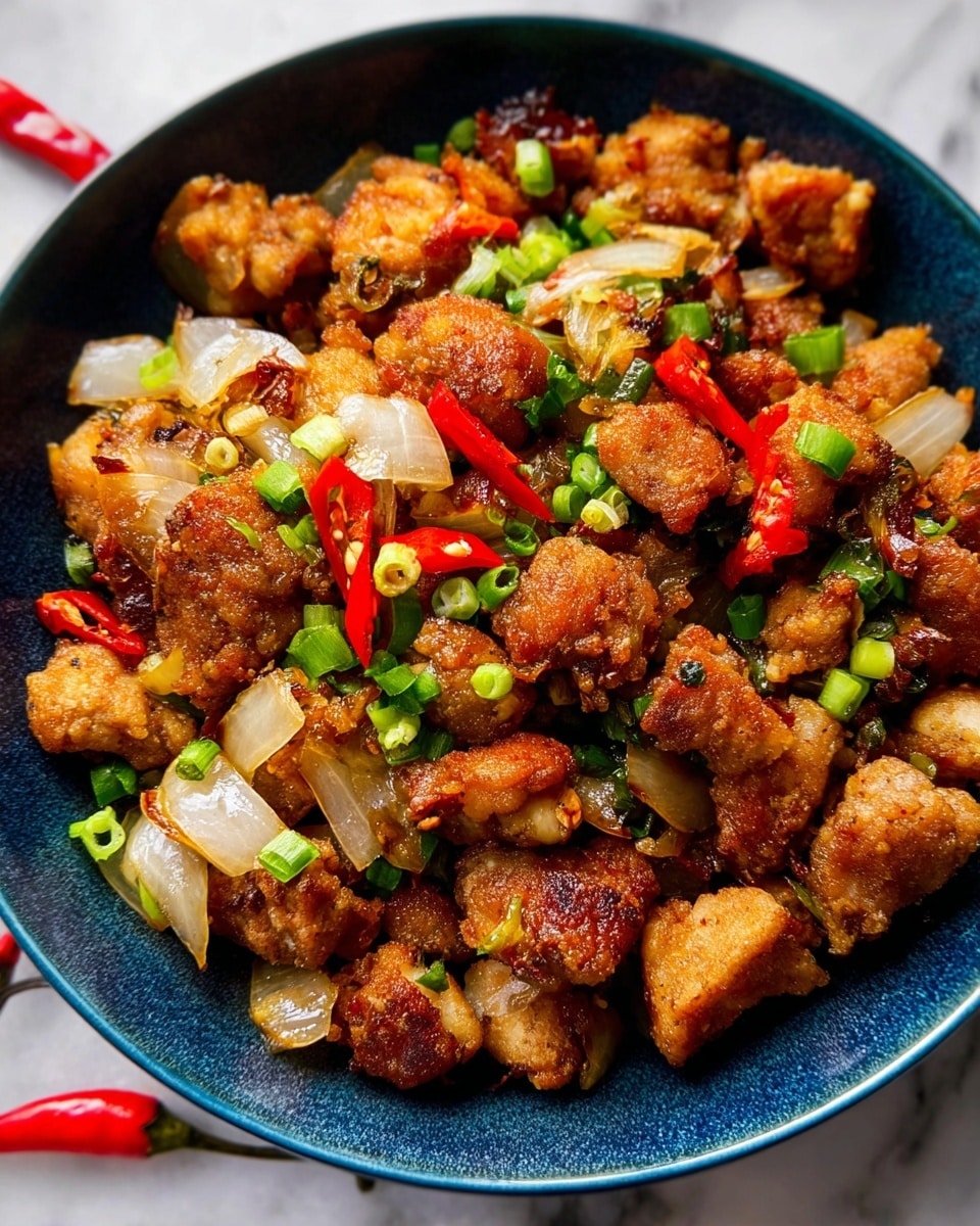 A dark blue bowl filled with many pieces of golden brown fried chicken chunks mixed with sautéed white onion pieces and small green and red chili slices, creating a colorful and textured dish. The chicken pieces have a crispy outside layer, surrounding the tender inside, while the onions add a glossy, soft contrast. Red chili pieces and green spring onions sit on top and throughout, adding bright spots of color. The bowl is on a surface with a white marbled texture. photo taken with an iphone --ar 4:5 --v 7
