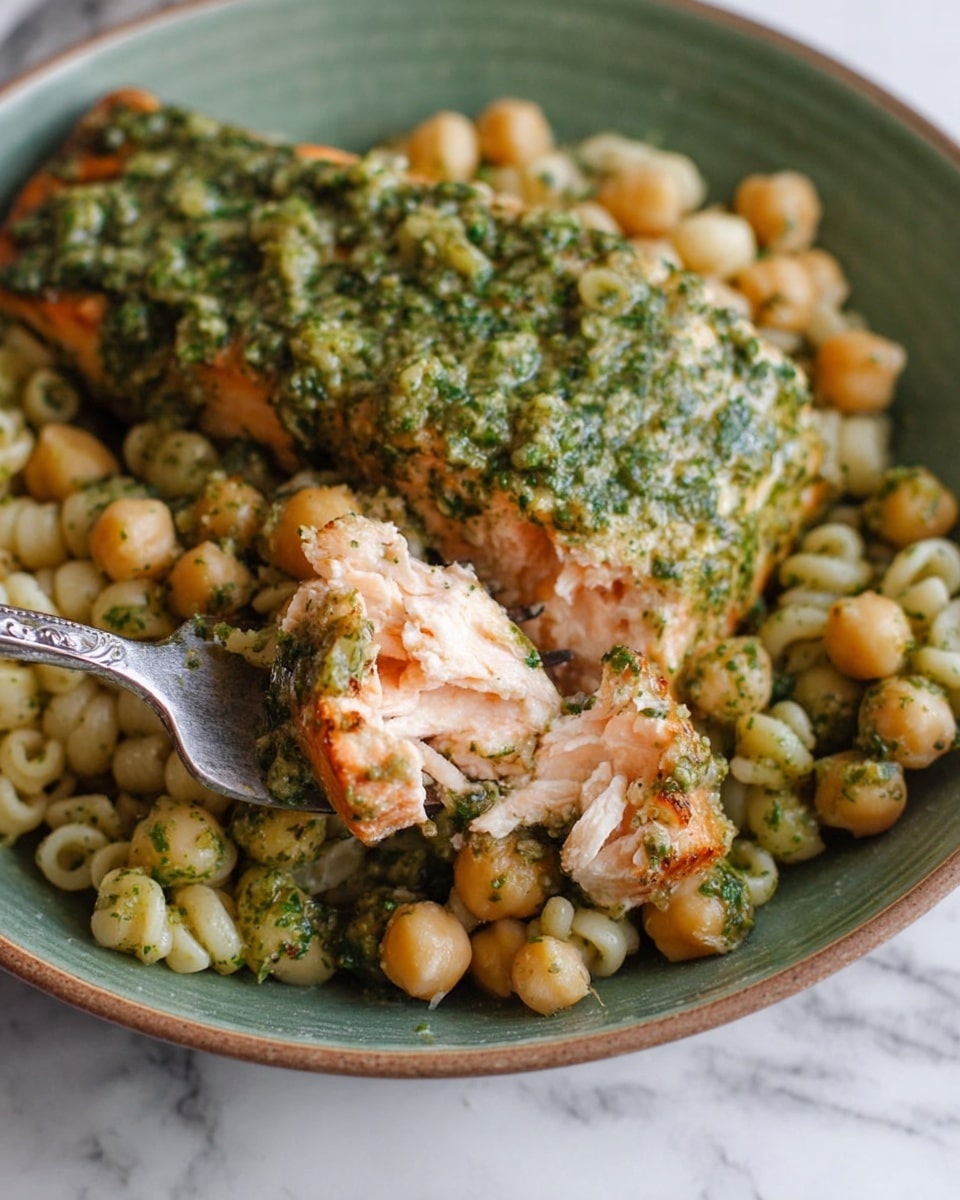 The image shows a close-up of a green bowl filled with two main layers: the bottom layer has small round pasta mixed with chickpeas, all lightly coated in a green herb sauce, giving it a slightly glossy and textured look, while the top layer features a thick piece of cooked salmon covered with more of the same green herb sauce, which looks rich and slightly chunky; a silver fork is lifting a piece of the salmon, revealing its flaky and moist pink interior, with some of the sauce clinging to the fish. The dish is set against a white marbled surface. photo taken with an iphone --ar 4:5 --v 7