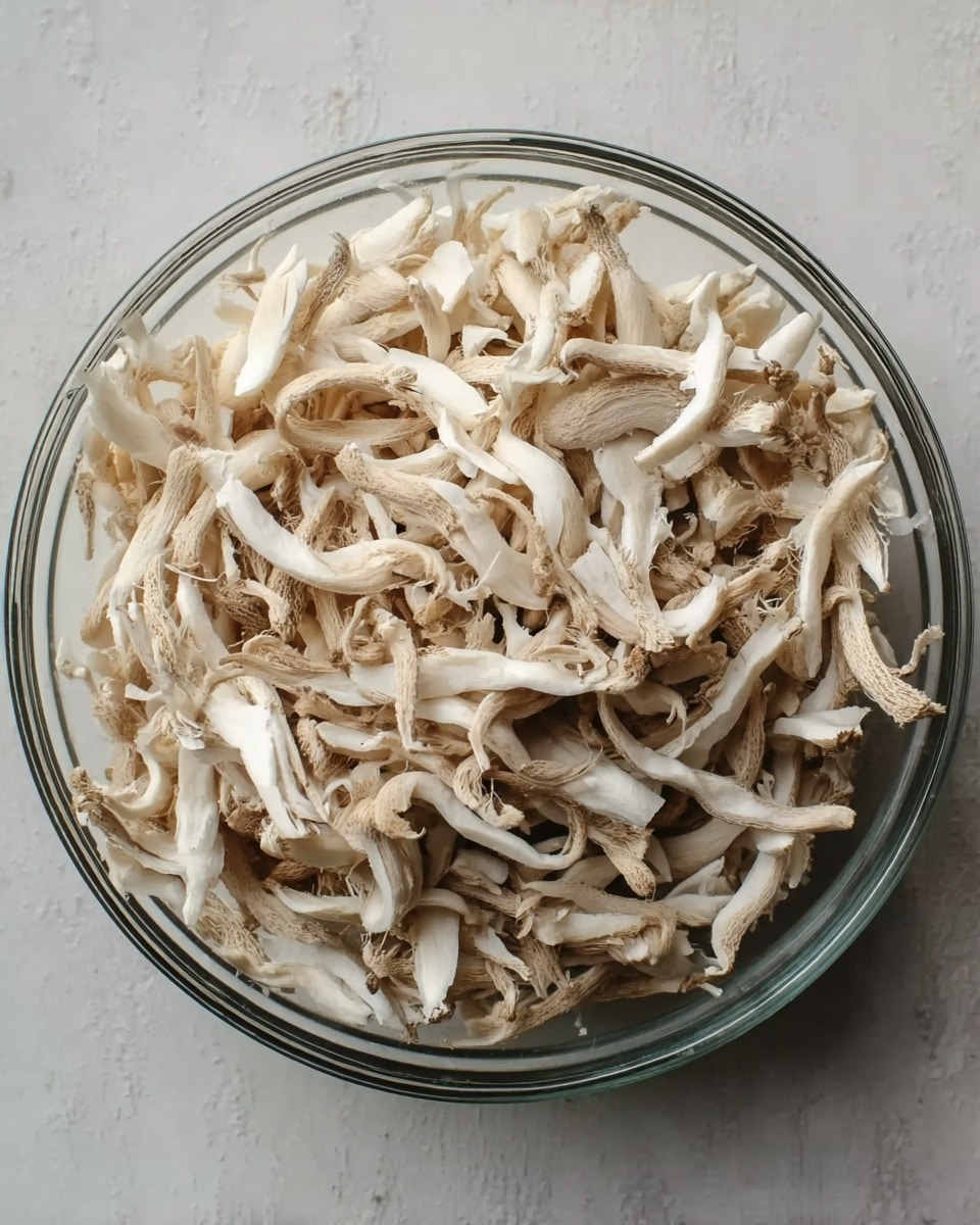 A clear round bowl filled with a large amount of shredded light beige mushroom pieces piled up evenly inside. The mushroom strips show a mix of thin and thick textures with some slight curves and frayed edges. The bowl is placed on a white marbled surface that highlights the neutral colors of the mushrooms. The overall visual is simple and natural, focusing on the texture and color variations of the mushroom strips. photo taken with an iphone --ar 4:5 --v 7