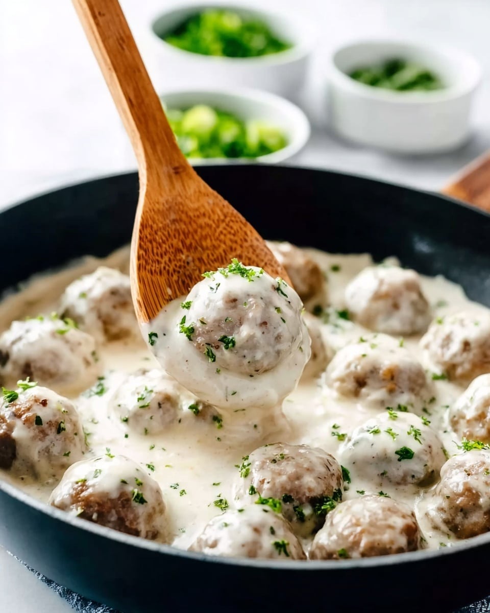 A black pan filled with round meatballs covered in thick, creamy white sauce scattered with small green herb bits. One woman's hand is holding a wooden spoon lifting a meatball coated in the sauce above the surface, showing texture with some small green herbs on it. In the background, blurred white bowls hold green chopped herbs and other ingredients, all set on a white marbled surface. photo taken with an iphone --ar 4:5 --v 7