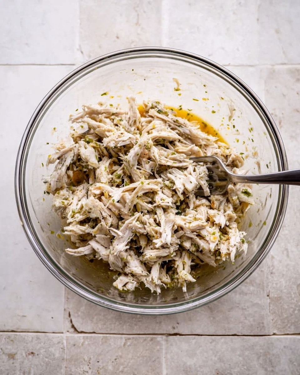 A clear glass bowl sits on a white marbled surface, filled with shredded white meat mixed with small green herb pieces and a light brown sauce at the bottom. The meat is piled loosely, showing a soft and fibrous texture. A silver fork rests inside the bowl, partially buried in the mixture, with the handle extending out to the left side. The overall colors are light with white and beige tones from the meat, dotted with small green bits from the herbs, and hints of golden brown from the sauce photo taken with an iphone --ar 4:5 --v 7