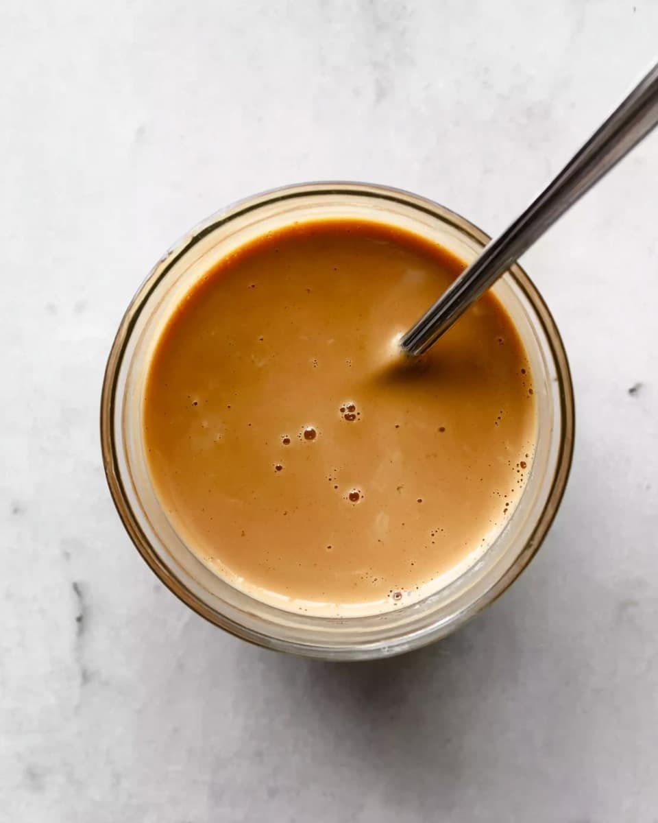 A clear glass bowl filled with a smooth, light brown sauce or liquid, showing a glossy texture on top with a few small bubbles. A thin, long metal spoon is placed inside the bowl, resting diagonally. The bowl sits on a white marbled surface which adds a subtle pattern around it. The photo taken with an iphone --ar 4:5 --v 7