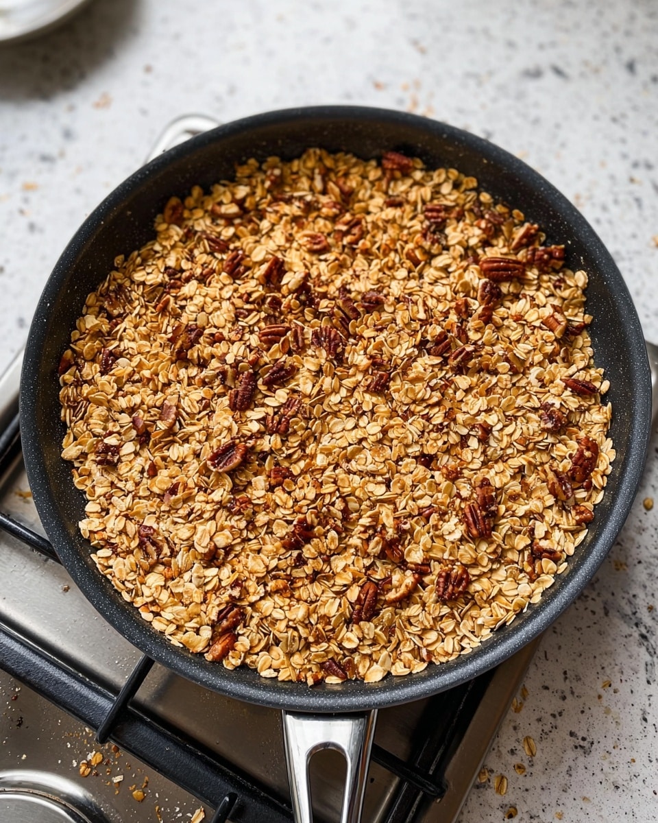 A black pan filled with a mix of toasted oats and pecan nuts, showing a golden brown color with pieces of pecans scattered evenly throughout. The pan sits on a stove burner, and the background is a white marbled surface with some small scattered oat flakes nearby. The texture looks dry and crunchy with a slightly rough surface from the oats and nuts combined photo taken with an iphone --ar 4:5 --v 7