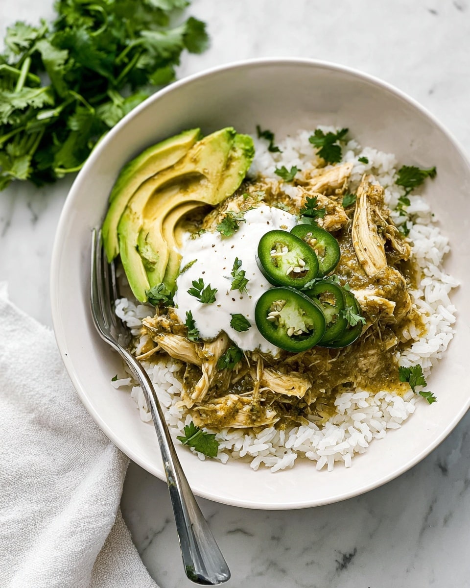 The dish shows a white bowl with three main layers: the bottom layer is white rice with a soft texture, the middle layer has shredded light brown chicken covered in a green sauce with a slightly chunky texture, and the top layer includes a dollop of white cream with two thin green slices of jalapeño and green cilantro leaves scattered around. On the side of the bowl, there are two slices of avocado in a pale green shade. A silver fork is placed inside the bowl on the left side. The bowl sits on a white marbled surface with a white plate of fresh cilantro leaves nearby. Photo taken with an iphone --ar 4:5 --v 7