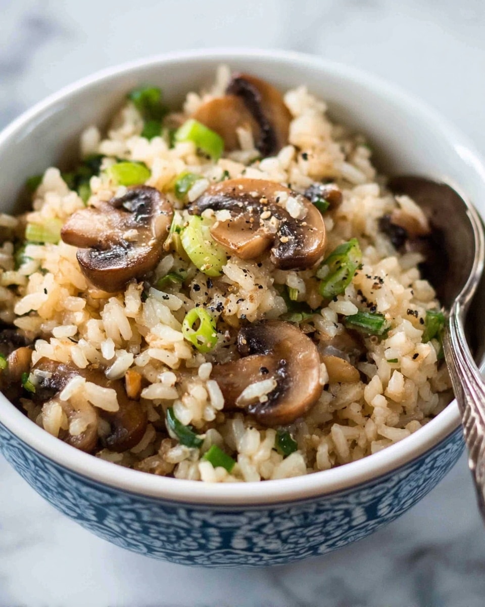 A close-up view of a bowl filled with cooked rice mixed with sliced brown mushrooms and small green onion pieces, with some black pepper sprinkled on top. The bowl is white with blue patterned detailing on the outside, and there is a silver spoon resting inside the bowl on the right side. The texture of the rice appears soft and the mushrooms have a slightly glossy surface. The background is a white marbled texture. Photo taken with an iphone --ar 4:5 --v 7