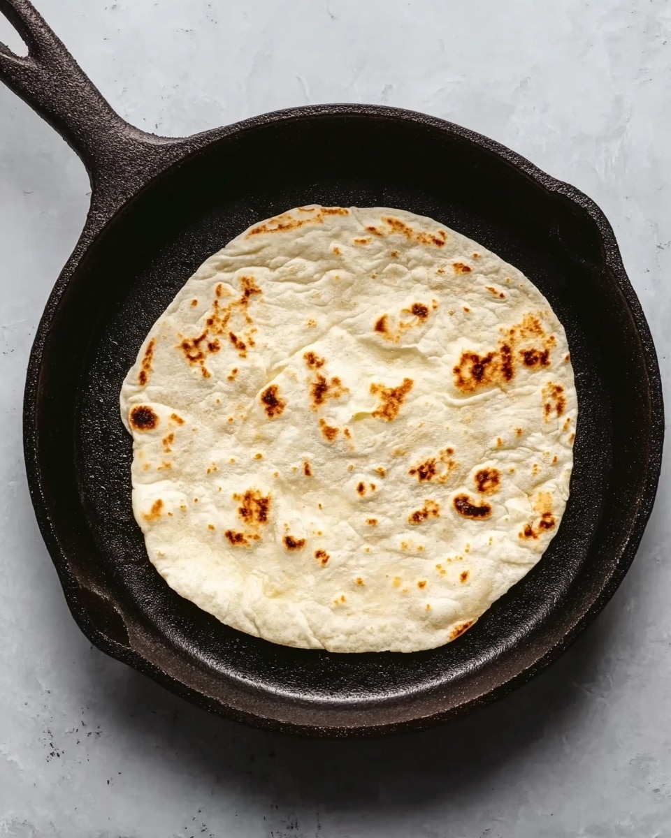 A single round flatbread with a light cream color and small brown spots is placed in the center of a black cast iron pan. The flatbread has slight uneven edges and a soft texture that shows some air bubbles. The background is a white marbled surface. photo taken with an iphone --ar 4:5 --v 7