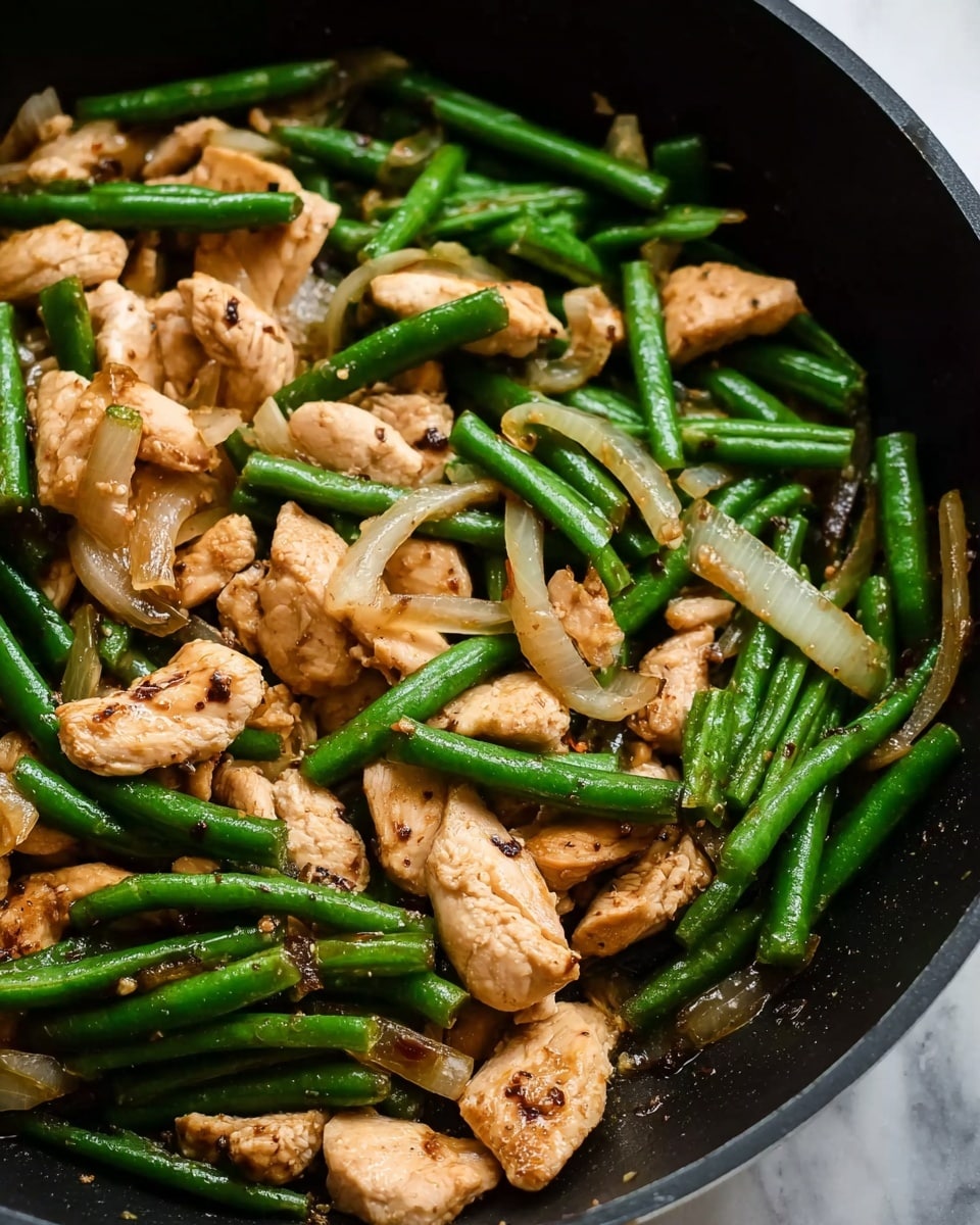 This image shows a close-up of a stir-fry dish in a black pan. The dish has three main layers: the bottom layer is light brown cooked chicken pieces with a slightly dry texture and some char marks, scattered evenly. The middle layer has bright green fresh green beans, cooked but still firm, mixed well with the chicken. The top layer includes translucent cooked white onion slices placed randomly over the chicken and beans. The background around the pan is a white marbled texture. photo taken with an iphone --ar 4:5 --v 7