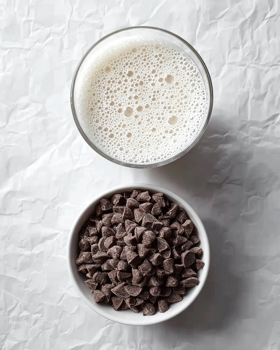 The image shows two round containers on a white marbled surface with crumpled paper texture around. The top container is a clear glass filled with a foamy white liquid, showing many small bubbles across the surface. Below it is a white bowl filled with small, dark brown chocolate chips, each with a smooth texture and somewhat shiny finish. The arrangement is simple, with the containers placed one above the other in the center of the frame. photo taken with an iphone --ar 4:5 --v 7