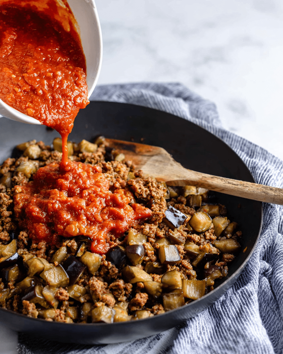 A black frying pan holds a cooked mix of small brown crumbled meat and medium chunks of light brown and purple eggplant pieces spread evenly. A wooden spoon rests in the pan, partially covered by the food. Above the pan, a white bowl tilted forward is pouring thick, bright red tomato sauce with a slightly chunky texture onto the meat and eggplant mix. The pan sits on a soft blue and white striped cloth over a white marbled surface. Photo taken with an iphone --ar 4:5 --v 7
