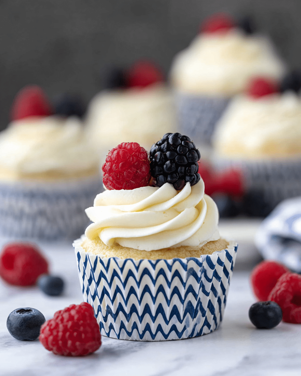 A cupcake in a white and blue zigzag patterned wrapper sits on a white marbled texture, topped with a swirl of white cream frosting. On top of the frosting, there is one red raspberry and one dark blackberry placed side by side. In the background, there are more cupcakes with the same frosting and berries, slightly blurred. Scattered around the base are loose raspberries and blackberries, adding color contrast. photo taken with an iphone --ar 4:5 --v 7