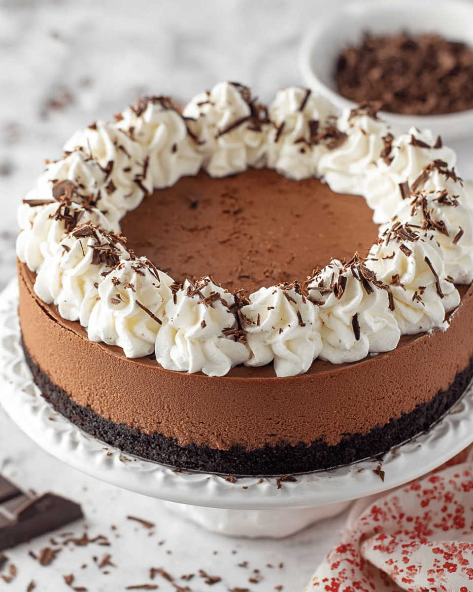 A round chocolate cheesecake with three visible layers: a dark brown crumbly crust at the bottom, a smooth medium brown chocolate cheesecake layer in the middle, and a ring of white whipped cream swirls on top around the edge. The whipped cream swirls are sprinkled with small thin chocolate shavings. The cake sits on a white scalloped cake stand. Around the cake are some scattered chocolate shavings, and a bowl of extra chocolate shavings is blurred in the white marbled background. Photo taken with an iphone --ar 4:5 --v 7