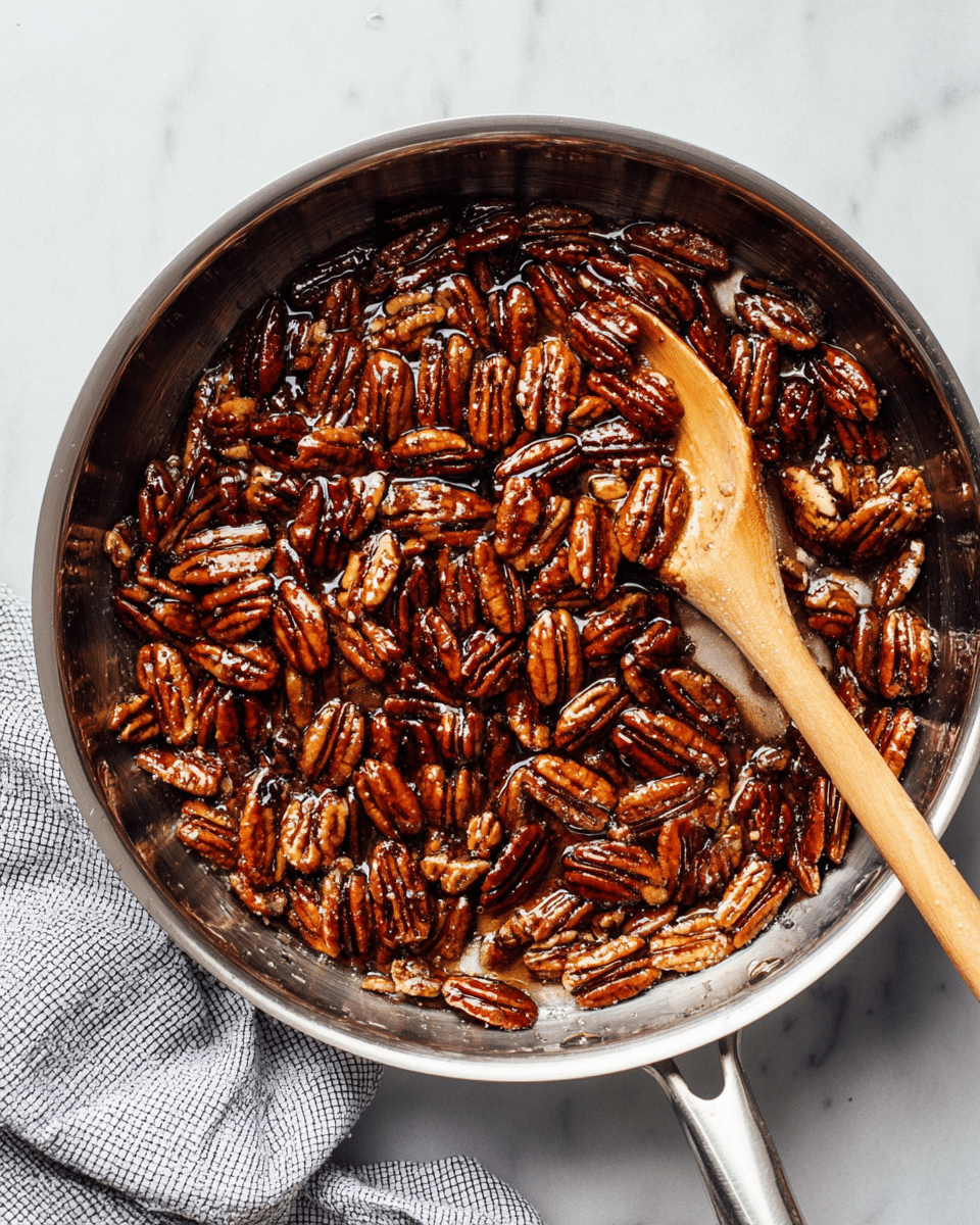 A shiny metal pan is filled with dark brown pecans coated in sticky syrup that looks shiny and thick. The pecans are spread evenly, covering the whole pan, and on the right side, a light wooden spoon with a smooth surface rests, partly covered in the syrup. The pan handle is wrapped with a cloth that has a white and black small check pattern. The pan sits on a white marbled surface, giving a clean and bright background. photo taken with an iphone --ar 4:5 --v 7