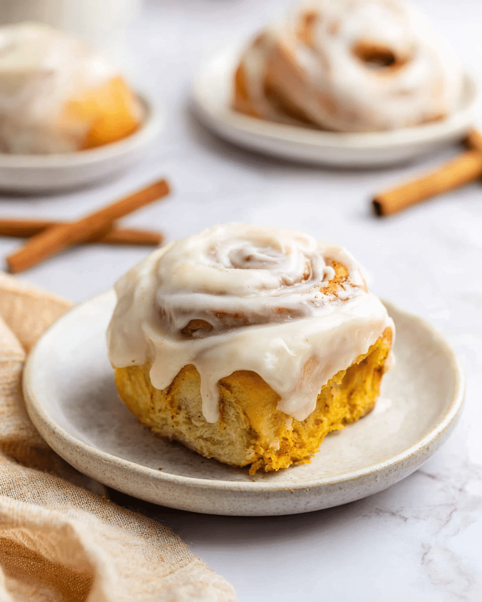 A single cinnamon roll sits on a small white plate placed on a white marbled surface. The roll shows two layers: a soft, golden-brown dough base with a slightly rough texture, topped with a thick layer of creamy white icing that swirls and drips slightly over the edges. In the background, there are two more white dishes holding cinnamon rolls with similar icing, softly blurred. A cinnamon stick and a beige cloth napkin are placed near the plate, enhancing the warm, cozy feeling. Photo taken with an iphone --ar 4:5 --v 7