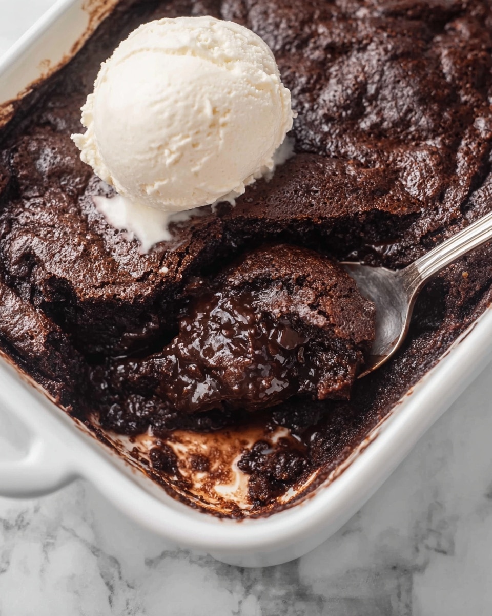 The image shows a white baking dish filled with a rich, dark brown chocolate pudding-like dessert with a soft and gooey texture. Inside the dish, a silver spoon scoops a portion of the thick dessert, revealing a gooey center with a slightly crusty top layer. A scoop of creamy, off-white vanilla ice cream sits on top of the dessert near the edge. The background is a white marbled surface, adding a clean and simple look. Photo taken with an iphone --ar 4:5 --v 7