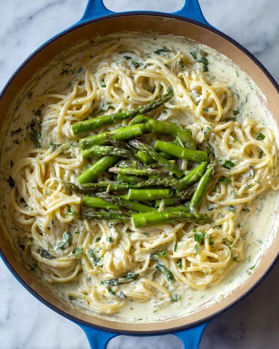 A round blue pot filled with creamy white pasta sauce mixed with spaghetti noodles that are twisted and spread evenly throughout the sauce, topped with bright green, thick asparagus pieces cut into short lengths and scattered in the center, all resting on a white marbled surface. The sauce has a smooth, rich texture with visible small green herb bits mixed within, coating the noodles well. Photo taken with an iphone --ar 4:5 --v 7