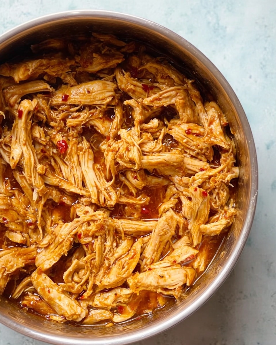 A metal bowl filled with shredded cooked chicken soaked in a reddish-brown sauce, with visible small bits of red chili or spices. The chicken pieces have a moist, fibrous texture and vary in size, mixed evenly in the sauce. The bowl sits on a white marbled surface, creating a clean and simple background. photo taken with an iphone --ar 4:5 --v 7