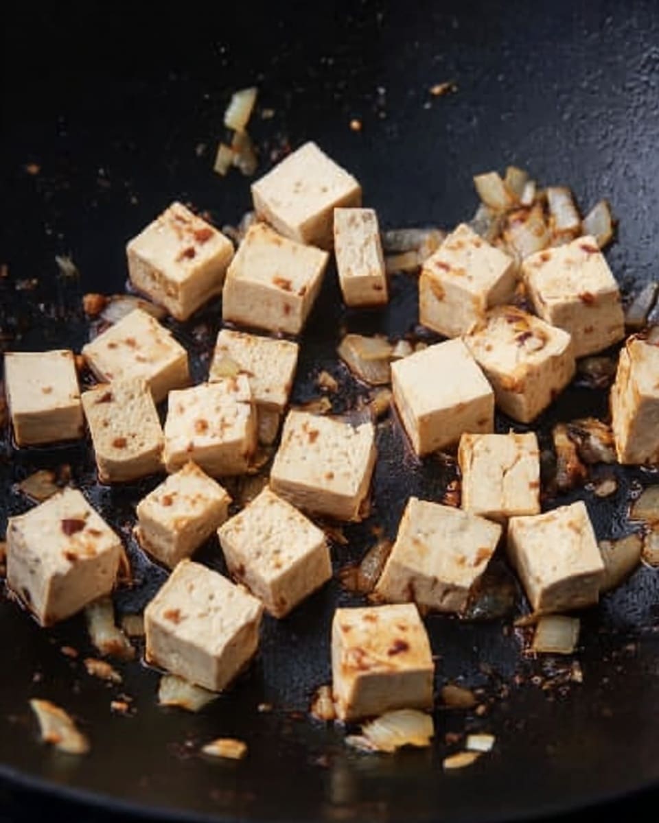 The image shows a black wok with small cubes of tofu being cooked. The tofu is light beige with some browned edges. There are small pieces of cooked onions, light golden and soft, scattered among the tofu cubes. The tofu and onions have a slight sheen from oil and some dark brown soy sauce spots. The wok’s surface has a slightly rough texture with some oil spots. Photo taken with an iphone --ar 4:5 --v 7