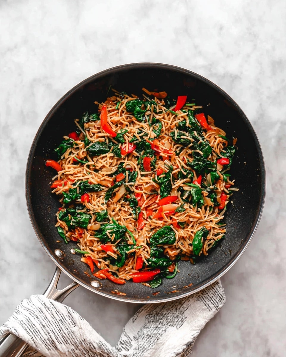 A black frying pan sits on a white marbled surface, filled with a colorful mix of cooked vegetables and noodles. The dish has one main layer of thin light brown noodles mixed with bright green spinach leaves and small red bell pepper pieces scattered evenly. The textures are soft and slightly wilted for the spinach, tender and lightly browned for the noodles, and firm for the bell peppers. The pan handle is wrapped with a white and gray striped kitchen cloth. Photo taken with an iphone --ar 4:5 --v 7