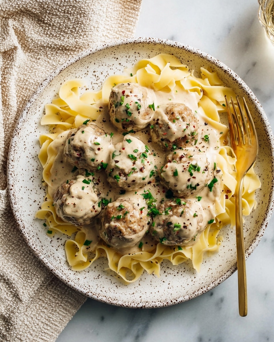 The image shows a white plate with speckled brown dots filled with pale yellow flat noodles as the bottom layer, topped with six round meatballs covered in a creamy beige sauce. The meatballs are sprinkled with small chopped green herbs and ground black pepper for texture. A shiny gold fork is placed on the right side of the plate, resting partly on the noodles. The plate sits on a white marbled surface next to a beige textured cloth. Photo taken with an iphone --ar 4:5 --v 7