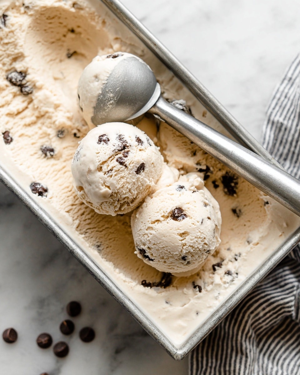 The image shows a close-up of a loaf pan filled with creamy light beige ice cream that has dark chocolate chips scattered throughout. Two round scoops of the ice cream sit on top, showing a smooth, slightly textured surface with visible chocolate chips embedded inside. The ice cream is soft and has a few uneven edges, indicating it has been recently scooped. A metal ice cream scoop, resting inside the pan, holds a small amount of the ice cream as if just used. The scene is set on a white marbled surface with a striped cloth partially visible nearby. photo taken with an iphone --ar 4:5 --v 7
