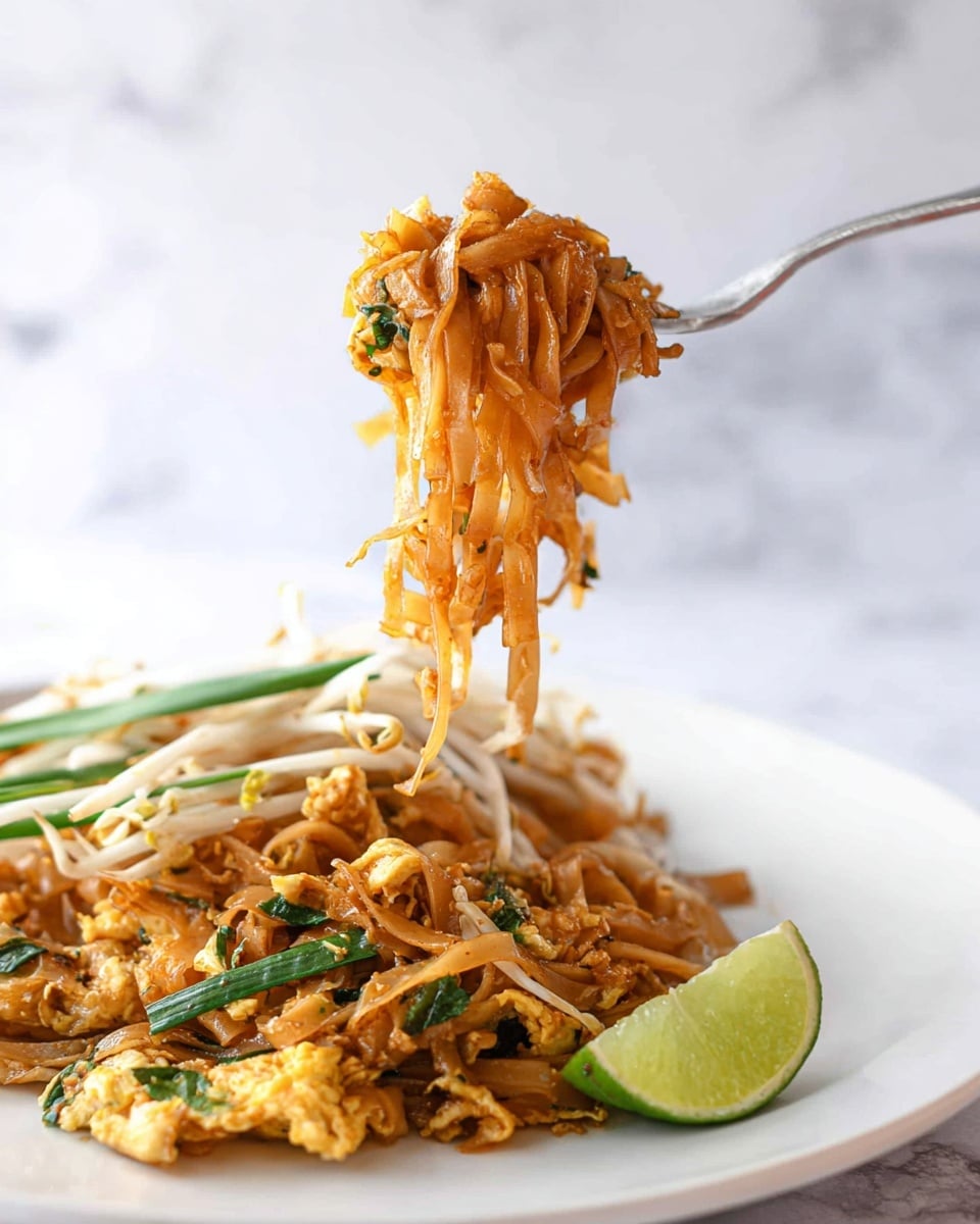 The image shows a fork holding up a twisted bundle of orange-brown noodles with some small green herb pieces mixed in. Below the fork is a white plate filled with layers of noodles mixed with cooked scrambled egg pieces, green onion stalks, and white bean sprouts. A lime wedge rests on the right side of the plate among the food. The background is a smooth white marbled surface with soft lighting. photo taken with an iphone --ar 4:5 --v 7