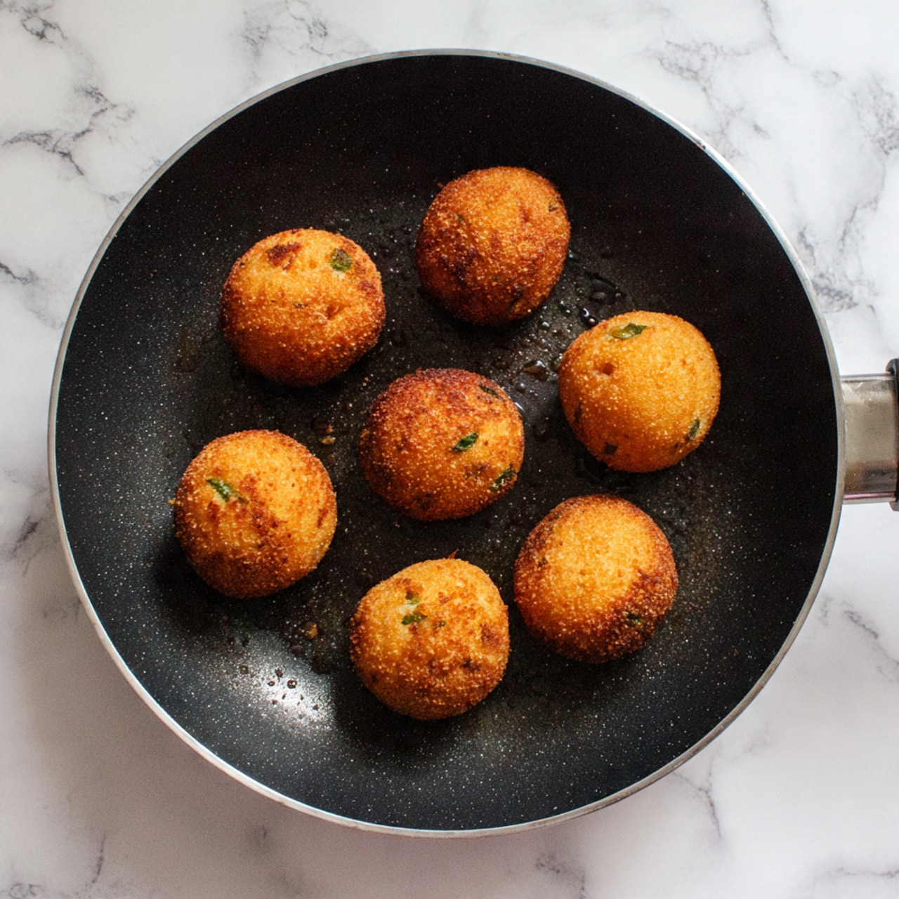 There are seven golden-brown round balls with a slightly rough texture and some small green bits inside, placed on a black pan with a slightly shiny surface. The pan rests on a white marbled surface, and the balls are spaced evenly in a circular pattern. The heating marks and browning make them look crispy on the outside. photo taken with an iphone --ar 4:5 --v 7