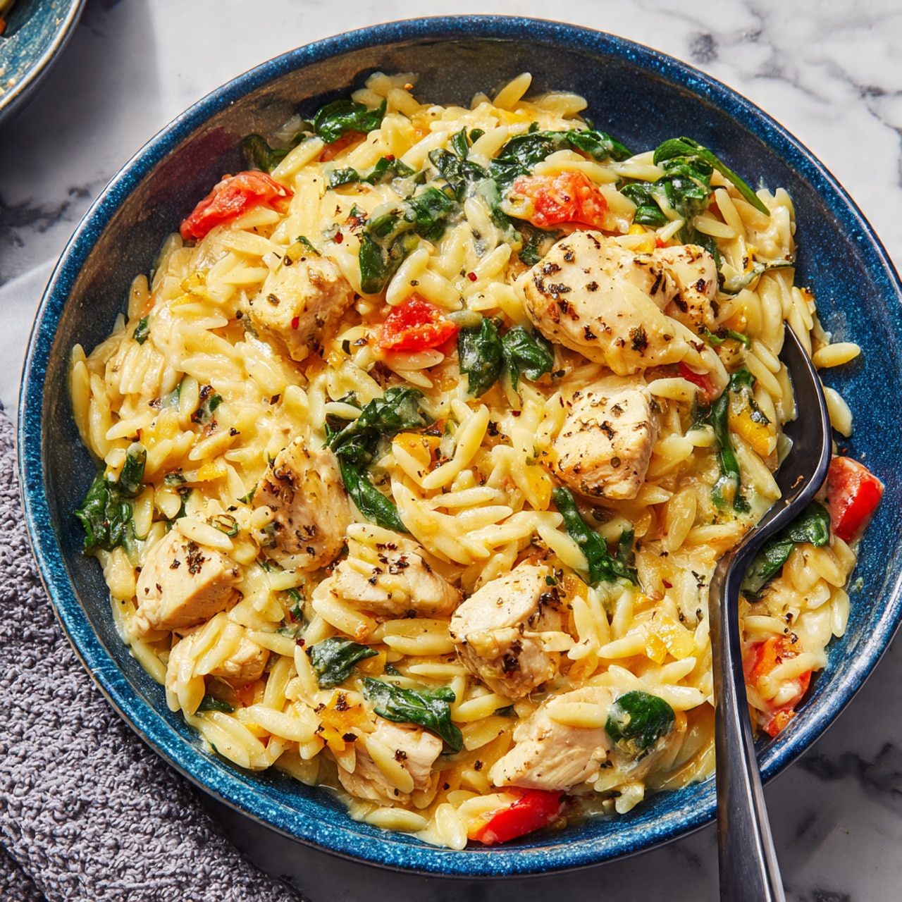 A large, shallow frying pan with a blue handle sits on a white marbled surface, filled with a creamy orange-colored rice dish made with small pasta-shaped grains, tender chunks of light brown chicken, and green leafy vegetables mixed throughout. There are small bits of red pepper or tomato scattered evenly, and a wooden spoon rests inside the pan, slightly stirring the food. The texture looks soft and rich, with a mix of starchy grains and pieces of chicken coated in the sauce. photo taken with an iphone --ar 4:5 --v 7