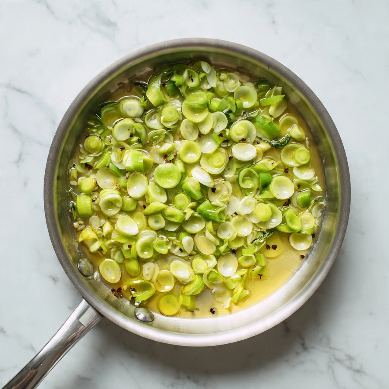 A metal pan sits on a white marbled surface, filled with small round green and white pieces of chopped vegetables that look like celery or leeks. The vegetables are floating and lightly cooked in clear oil or butter, covering the bottom of the pan evenly. The pan handle extends out to the left side of the image. Photo taken with an iphone --ar 4:5 --v 7