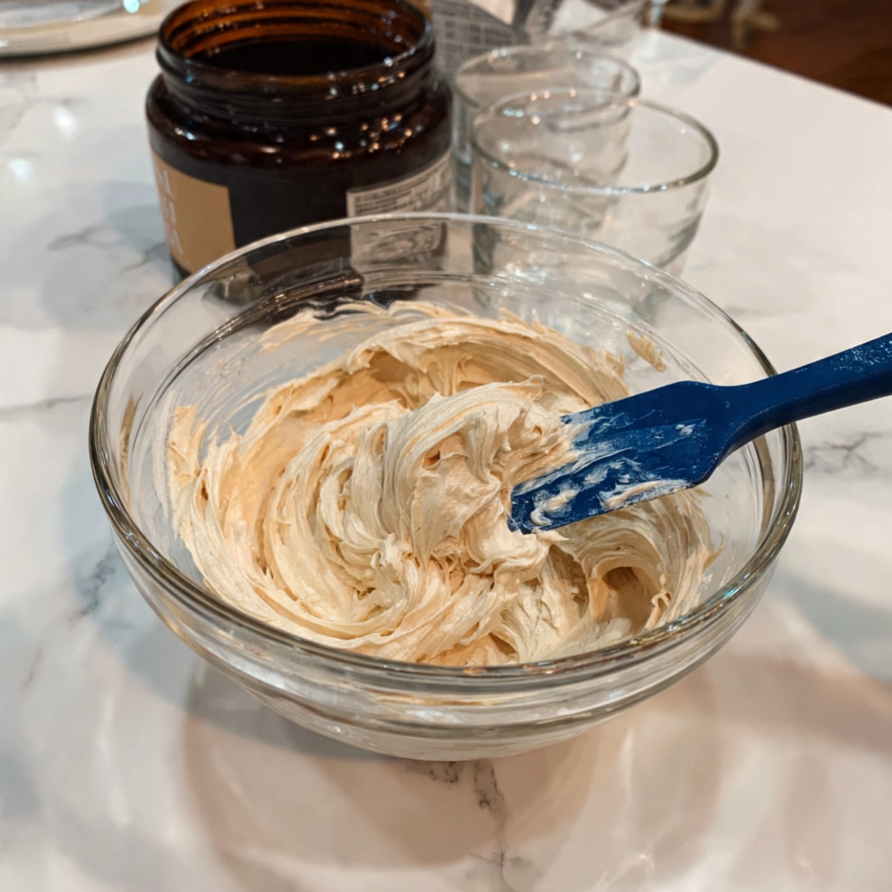 A clear glass bowl filled with one thick layer of light beige whipped cream-like mixture being stirred with a blue spatula, showing smooth, soft, and fluffy texture with swirls and peaks. Behind the bowl, there is a dark brown container and a clear glass cup on a white marbled surface. The photo taken with an iphone --ar 4:5 --v 7