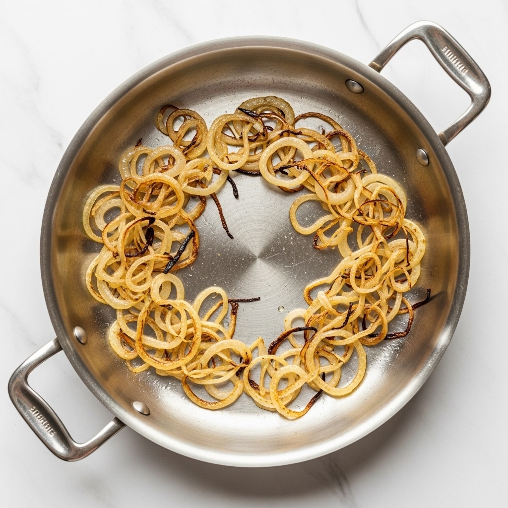 A round stainless steel pan with two handles shows thinly cooked caramelized onions spread unevenly in the center, forming a loose ring. The onions are golden brown with darker spots and look soft and slightly translucent, sitting against the shiny silver surface of the pan. The background is a white marbled texture. Photo taken with an iphone --ar 4:5 --v 7