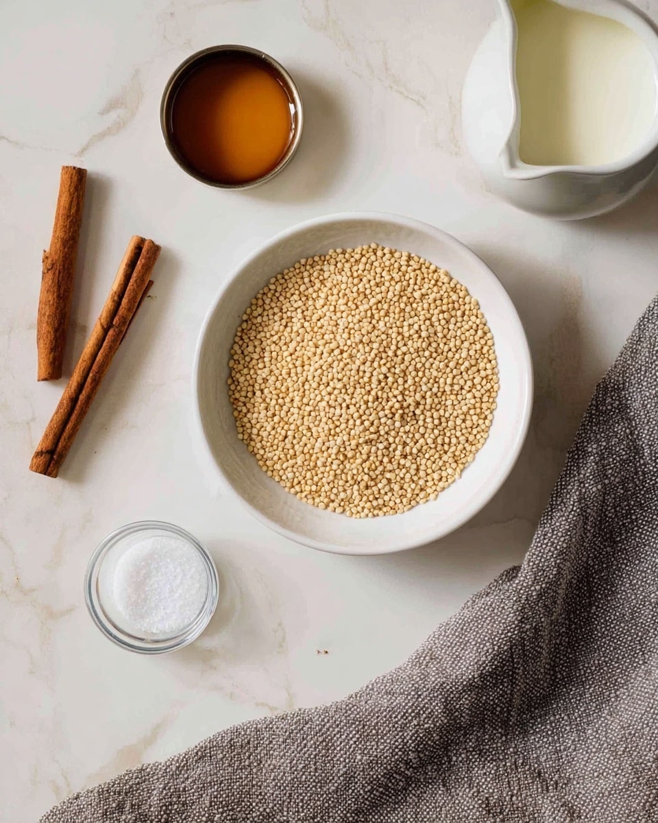 The image shows a white bowl filled with small, round, light beige grains placed on a white marbled surface. Around the bowl, there is a round glass container with white salt in the front left, two light brown cinnamon sticks on the left side, a small metal cup with golden honey above the cinnamon sticks, and a white small pitcher with creamy white milk at the top right. A textured gray cloth is spread on the right side, partially in the frame. Photo taken with an iphone --ar 4:5 --v 7
