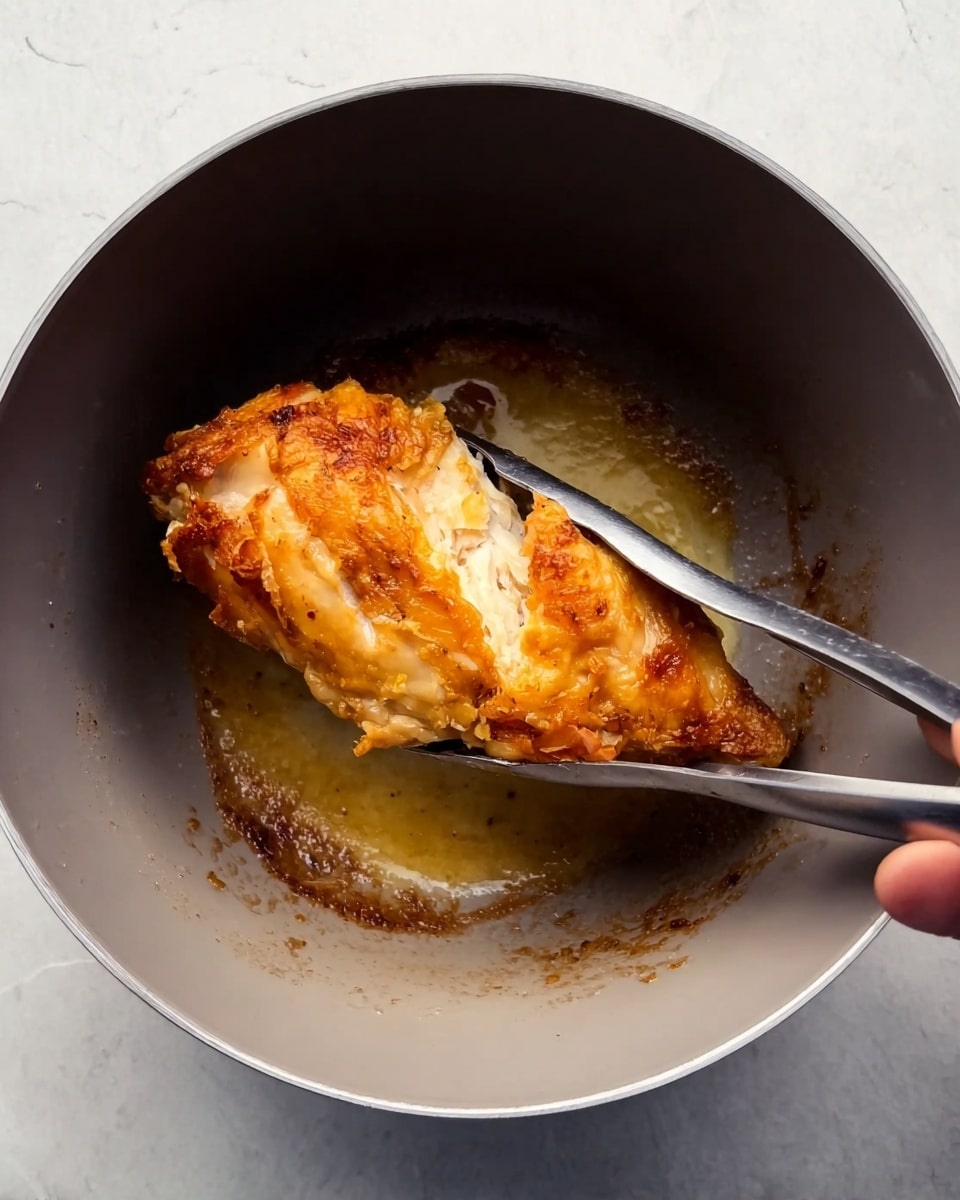A close-up image shows a piece of golden brown cooked chicken being held with silver tongs inside a deep grey pot. The chicken has a crispy-looking outer layer with some lighter, softer parts visible on the surface. The inside of the pot shows some browned oil or cooking residue, giving a slightly textured base beneath the chicken. A woman's hand is holding the silver tongs on the right side of the frame. The whole scene is set against a white marbled textured background. Photo taken with an iphone --ar 4:5 --v 7