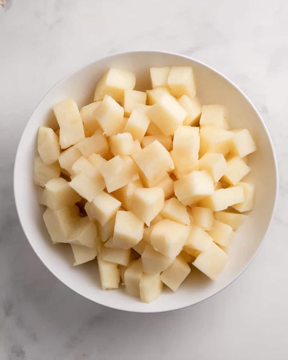 A white bowl filled with many small cubes of peeled potato, all light beige in color, sitting on a white marbled surface. The potato cubes are neatly piled up filling the bowl almost to the top, showing a soft, smooth texture. There are no other ingredients or decorations in the bowl or around it, keeping the focus on the diced potatoes. Photo taken with an iphone --ar 4:5 --v 7
