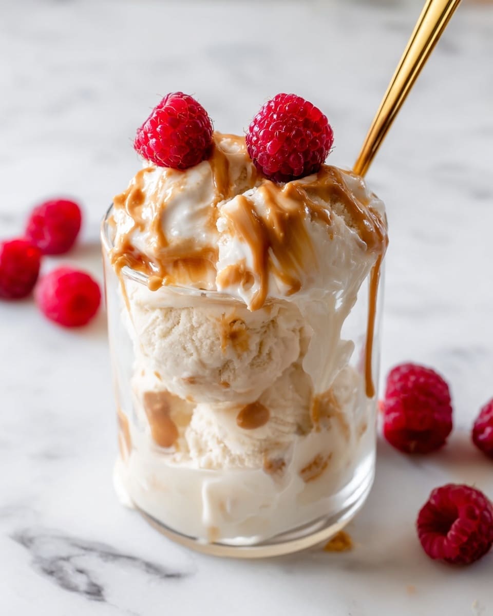 This image shows a clear glass filled with three scoops of creamy white ice cream. On top of the ice cream are swirls of light brown peanut butter and three bright red raspberries placed near the center. The ice cream is melting slightly, creating soft white drips down the sides of the glass. A gold spoon is inserted into the ice cream from the back right. The glass sits on a white marbled surface with a few raspberries scattered around it. Photo taken with an iphone --ar 4:5 --v 7