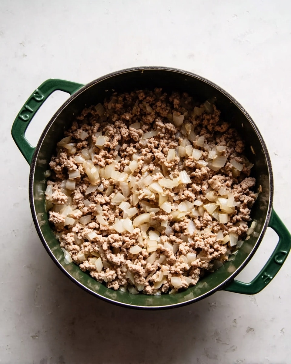 A close-up top view of cooked ground meat mixed with translucent, soft, cooked onion pieces in a dark green cooking pot with two handles, resting on a white marbled surface. The meat is light brown with small crumb-like texture, while the onion pieces are scattered evenly throughout, showing a slightly glossy look from cooking. The pot is centered with no other items around it, creating a simple and clear focus on the mixture inside. photo taken with an iphone --ar 4:5 --v 7