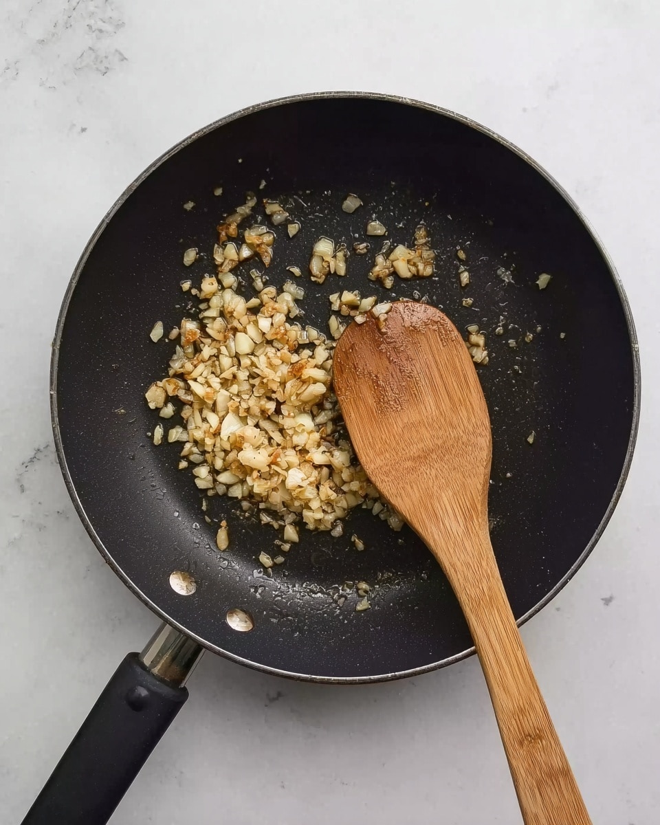 A black frying pan with a smooth surface holds finely chopped garlic that is lightly browned and sizzling. A wooden spoon with a black handle rests inside the pan, slightly tilted and mixing the garlic. The background is a white marbled texture. photo taken with an iphone --ar 4:5 --v 7