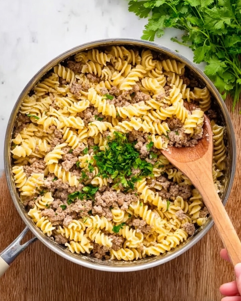 A large metal pan filled with spiral pasta mixed with cooked ground meat, evenly spread throughout. The pasta is light yellow with a smooth texture, and the meat is brown and crumbly, sitting mostly on top and between the pasta spirals. A woman's hand is holding a wooden spoon scooping some pasta and meat from the pan. Fresh green parsley leaves are sprinkled in the center on top of the pasta. The pan is placed on a white marbled surface with a bunch of fresh coriander leaves at the top right corner. Photo taken with an iphone --ar 4:5 --v 7