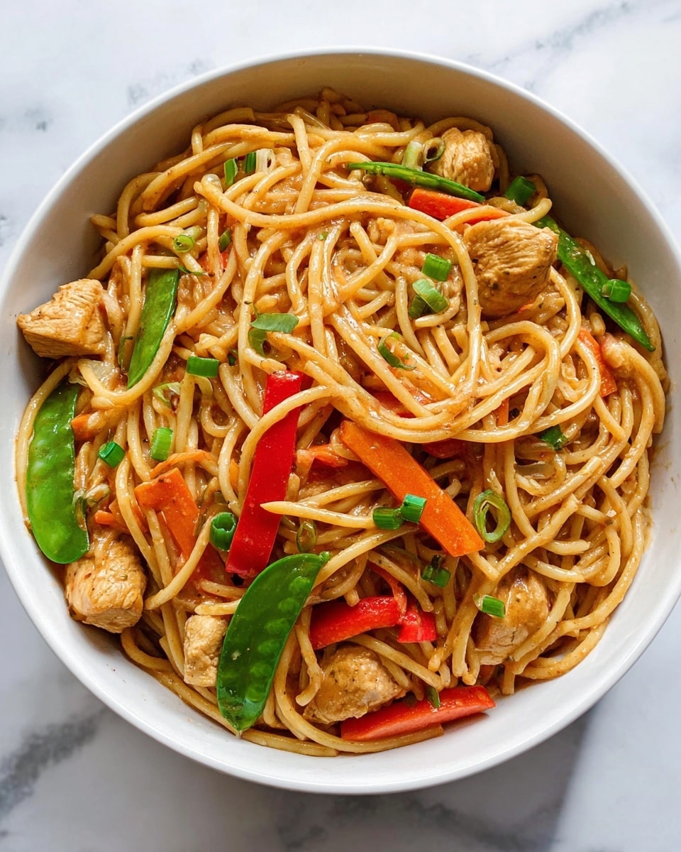 A white bowl filled with cooked spaghetti noodles coated in a light brown sauce, mixed with pieces of light tan chicken, bright orange carrot sticks, green snow peas, and red bell pepper slices. Small green garnish pieces are sprinkled on top. The bowl sits on a white marbled surface. Photo taken with an iphone --ar 4:5 --v 7