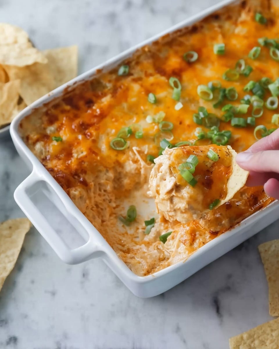 The dish is a baked casserole in a white ceramic rectangular dish. It has two main layers: a melted, golden-brown cheesy top layer with some green onion slices scattered on it, and a thick layer underneath with creamy, orange-tinted sauce mixed with rice and shredded chicken. A woman’s hand holds a scoop of the casserole with a soft tortilla chip to serve. The dish sits on a white marbled surface, with some tortilla chips visible nearby. Photo taken with an iphone --ar 4:5 --v 7