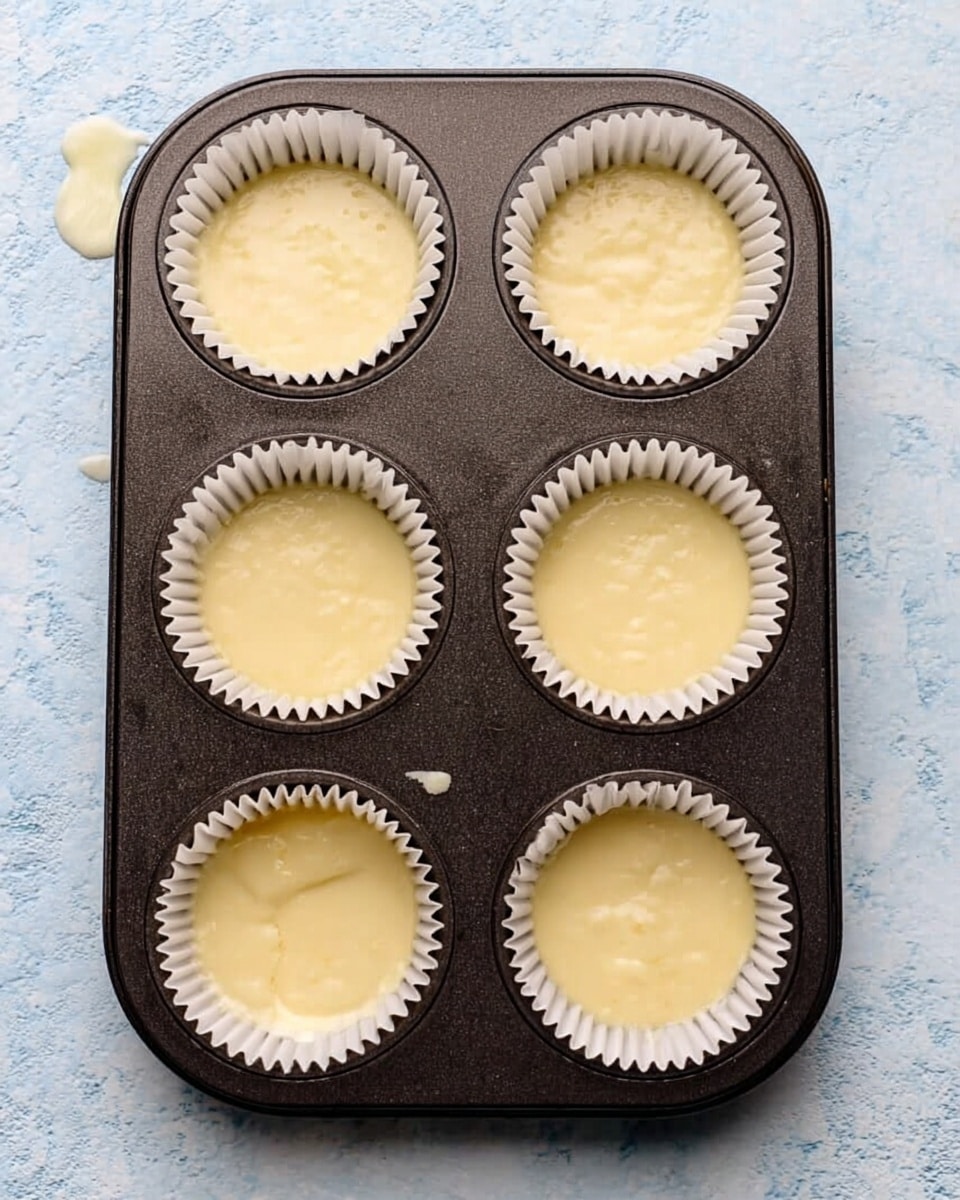 A dark metal muffin tray with six cups lined with white paper liners filled with smooth, pale yellow batter. The batter fills each liner about three-quarters full, showing a creamy and slightly shiny surface with small air bubbles. The tray sits on a white marbled surface with a faint texture, and a small drip of batter is visible on the left side near the edge of the tray. photo taken with an iphone --ar 4:5 --v 7