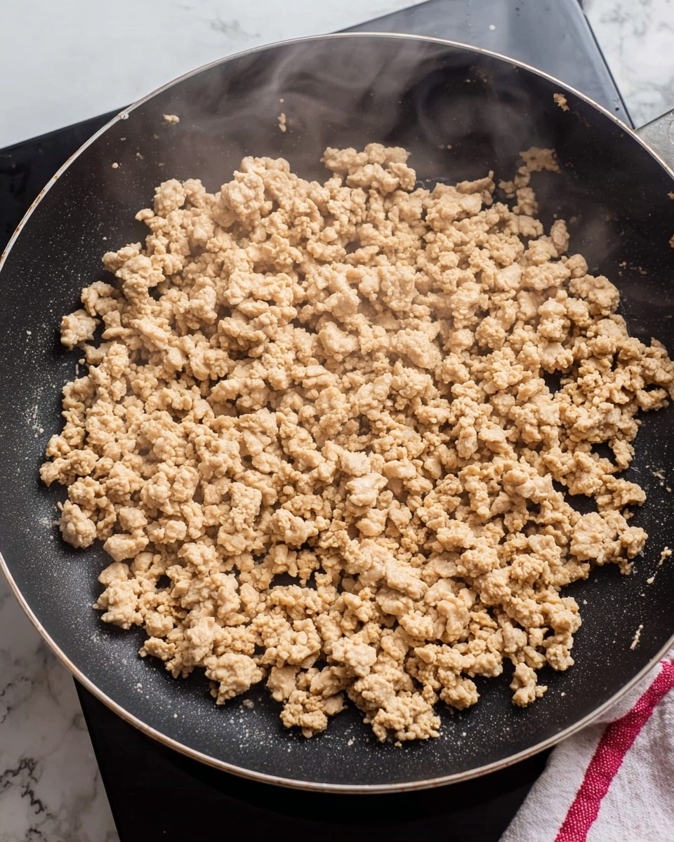 A close-up view of small, light beige crumbles of cooked ground meat spread evenly across the surface of a black non-stick frying pan. Wisps of steam rise gently from the pan, showing the heat of the freshly cooked food. The pan rests on a black stovetop with a white marbled texture surface visible at the corner, and a white and red striped cloth can be seen in the background. photo taken with an iphone --ar 4:5 --v 7