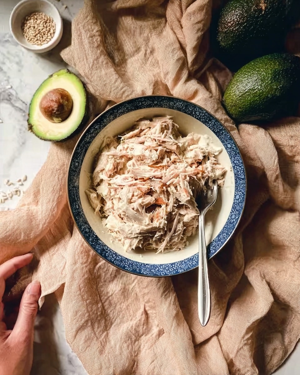 The image shows a blue and white bowl filled with shredded light beige tuna mixed with a creamy sauce, with some visible brown flakes on top, placed slightly off-center on a crumpled beige cloth over a white marbled surface. A silver fork is resting inside the bowl on the right side. Around the bowl, there are three whole dark green avocados and a small white bowl with light-colored seeds nearby. The scene is lit softly, emphasizing textures and colors, with a woman's hand barely touching the edge of the bowl. Photo taken with an iphone --ar 4:5 --v 7