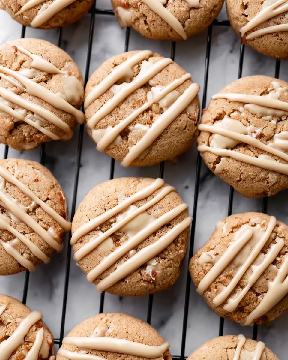 The image shows a grid of round baked cookies with a light brown, slightly cracked surface. Each cookie is decorated with a creamy beige drizzle that forms loose lines crossing over the top. The cookies are placed closely together on a black wire cooling rack, which is set against a white marbled background. The texture of the cookies looks soft with a few small bumps, and the drizzle adds a smooth, glossy layer on top. The photo taken with an iphone --ar 4:5 --v 7