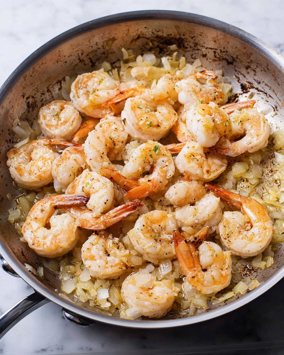 In a silver pan on a white marbled surface, there is one main layer of cooked shrimp and small chopped onions. The shrimp are light pink with some slightly browned spots and have an orange tail. The onions are soft, pale yellow, and spread evenly under and around the shrimp. The pan shows brown bits on the bottom, indicating cooking marks. photo taken with an iphone --ar 4:5 --v 7